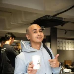 Raditya Dika berjaket abu-abu memegang gelas kopi di balik meja bar sebuah kafe