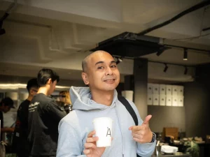 Raditya Dika berjaket abu-abu memegang gelas kopi di balik meja bar sebuah kafe