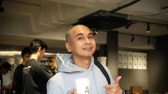 Raditya Dika berjaket abu-abu memegang gelas kopi di balik meja bar sebuah kafe