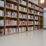 Deretan rak kayu berisi buku di sebuah perpustakaan universitas dengan lantai terang di depannya