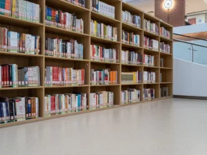 Deretan rak kayu berisi buku di sebuah perpustakaan universitas dengan lantai terang di depannya