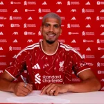 Ronald Araujo tersenyum mengenakan jersey merah Liverpool di depan dinding sponsor klub