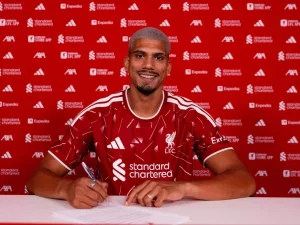Ronald Araujo tersenyum mengenakan jersey merah Liverpool di depan dinding sponsor klub