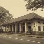 Ilustrasi bangunan rumah sakit bergaya Indis di tepi jalan lengang