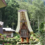Rumah adat tongkonan dan lumbung padi Toraja dengan dinding berukir geometris berwarna merah, hitam, putih, dan kuning di lereng berhutan
