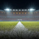 Rumput lapangan sepak bola dengan garis putih di bawah lampu sorot stadion pada malam hari, tribun penonton tampak buram di latar belakang