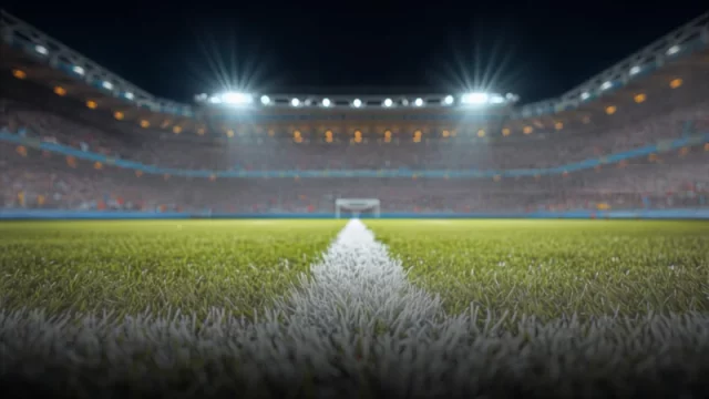 Rumput lapangan sepak bola dengan garis putih di bawah lampu sorot stadion pada malam hari, tribun penonton tampak buram di latar belakang