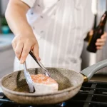 Ilustrasi wajan antilengket berlapis gelap di atas kompor dapur rumah tangga dengan potongan ikan yang sedang dimasak