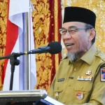 Seorang pejabat berseragam dinas berbicara di depan mikrofon dengan latar kain bermotif emas