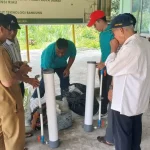 Sejumlah pria berdiri mengelilingi tiga tabung filtrasi putih di halaman berlatar spanduk kegiatan.
