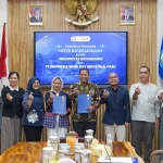 Sembilan orang berdiri berjajar memegang map biru di depan layar bertuliskan penandatanganan nota kesepahaman.