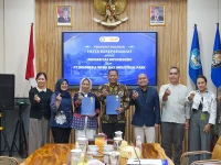 Sembilan orang berdiri berjajar memegang map biru di depan layar bertuliskan penandatanganan nota kesepahaman.