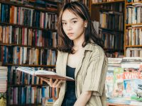 Ilustrasi seorang remaja perempuan membaca buku di antara rak toko buku.