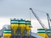 Deretan silo tinggi dan sebuah derek berdiri di kompleks pabrik semen di bawah langit terang