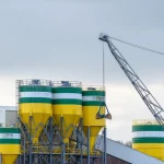 Deretan silo tinggi dan sebuah derek berdiri di kompleks pabrik semen di bawah langit terang