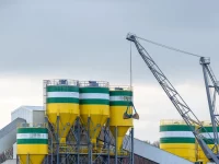 Deretan silo tinggi dan sebuah derek berdiri di kompleks pabrik semen di bawah langit terang