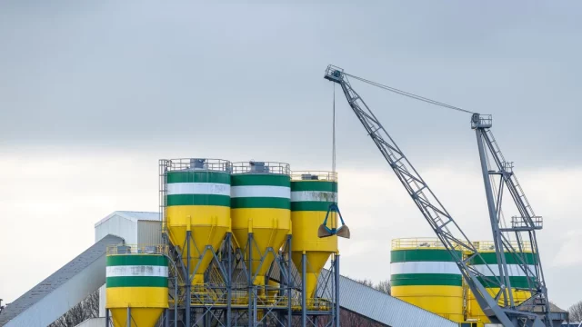 Deretan silo tinggi dan sebuah derek berdiri di kompleks pabrik semen di bawah langit terang