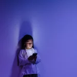 Seorang anak menatap layar ponsel yang menyala di ruangan berpencahayaan biru