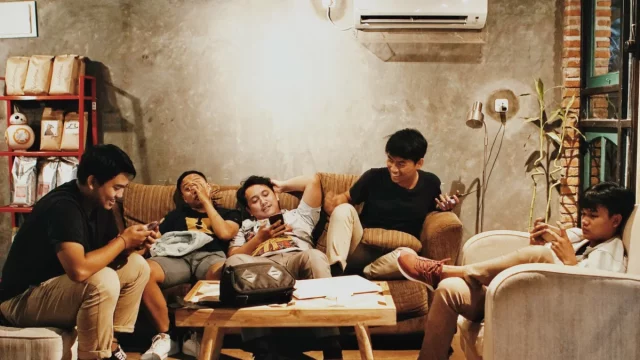 Enam anak muda duduk bersantai di sofa kafe sambil memegang ponsel