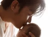 Ayah Asia berkaus putih mencium kening bayi yang digendongnya di dekat jendela terang
