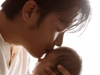 Ayah Asia berkaus putih mencium kening bayi yang digendongnya di dekat jendela terang