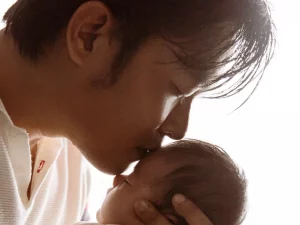 Ayah Asia berkaus putih mencium kening bayi yang digendongnya di dekat jendela terang