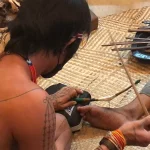 Seorang sikerei bertato sedang membuat tato Mentawai di kaki seseorang, di atas tikar anyaman