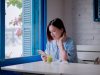 Ilustrasi seorang perempuan muda duduk di meja dekat jendela berdaun biru sambil menatap layar telepon pintarnya.