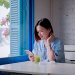Ilustrasi seorang perempuan muda duduk di meja dekat jendela berdaun biru sambil menatap layar telepon pintarnya.