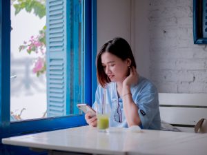 Ilustrasi seorang perempuan muda duduk di meja dekat jendela berdaun biru sambil menatap layar telepon pintarnya.