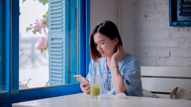 Ilustrasi seorang perempuan muda duduk di meja dekat jendela berdaun biru sambil menatap layar telepon pintarnya.