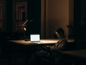 Ilustrasi seseorang menghadap layar laptop di ruangan yang temaram.
