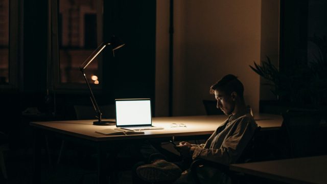 Ilustrasi seseorang menghadap layar laptop di ruangan yang temaram.