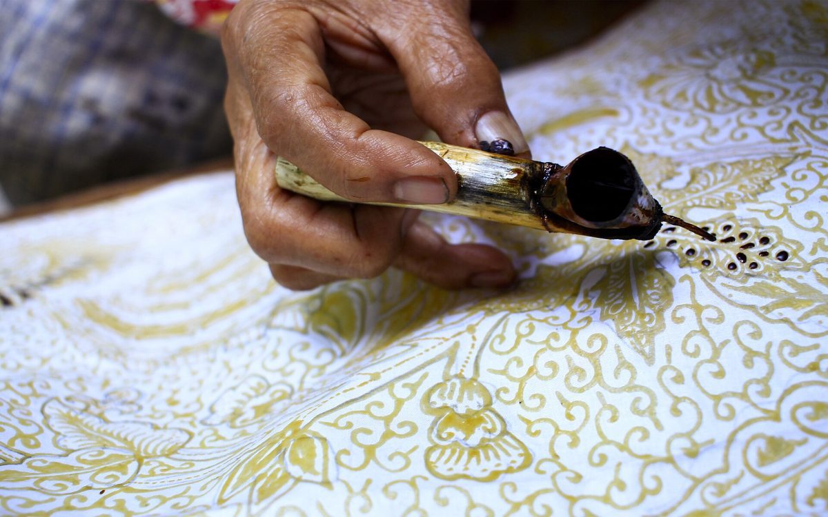 Ilustrasi tangan pembatik menorehkan malam dengan canting di atas kain bermotif.
