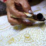 Ilustrasi tangan pembatik menorehkan malam dengan canting di atas kain bermotif.