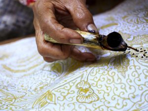 Ilustrasi tangan pembatik menorehkan malam dengan canting di atas kain bermotif.