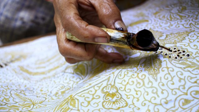 Ilustrasi tangan pembatik menorehkan malam dengan canting di atas kain bermotif.