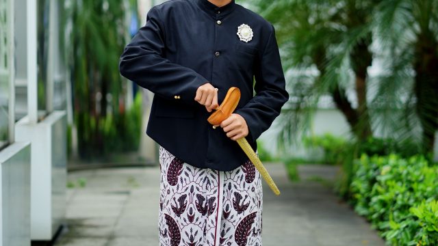 Ilustrasi seorang lelaki berbusana tradisi Jawa berdiri di halaman berpepohonan.