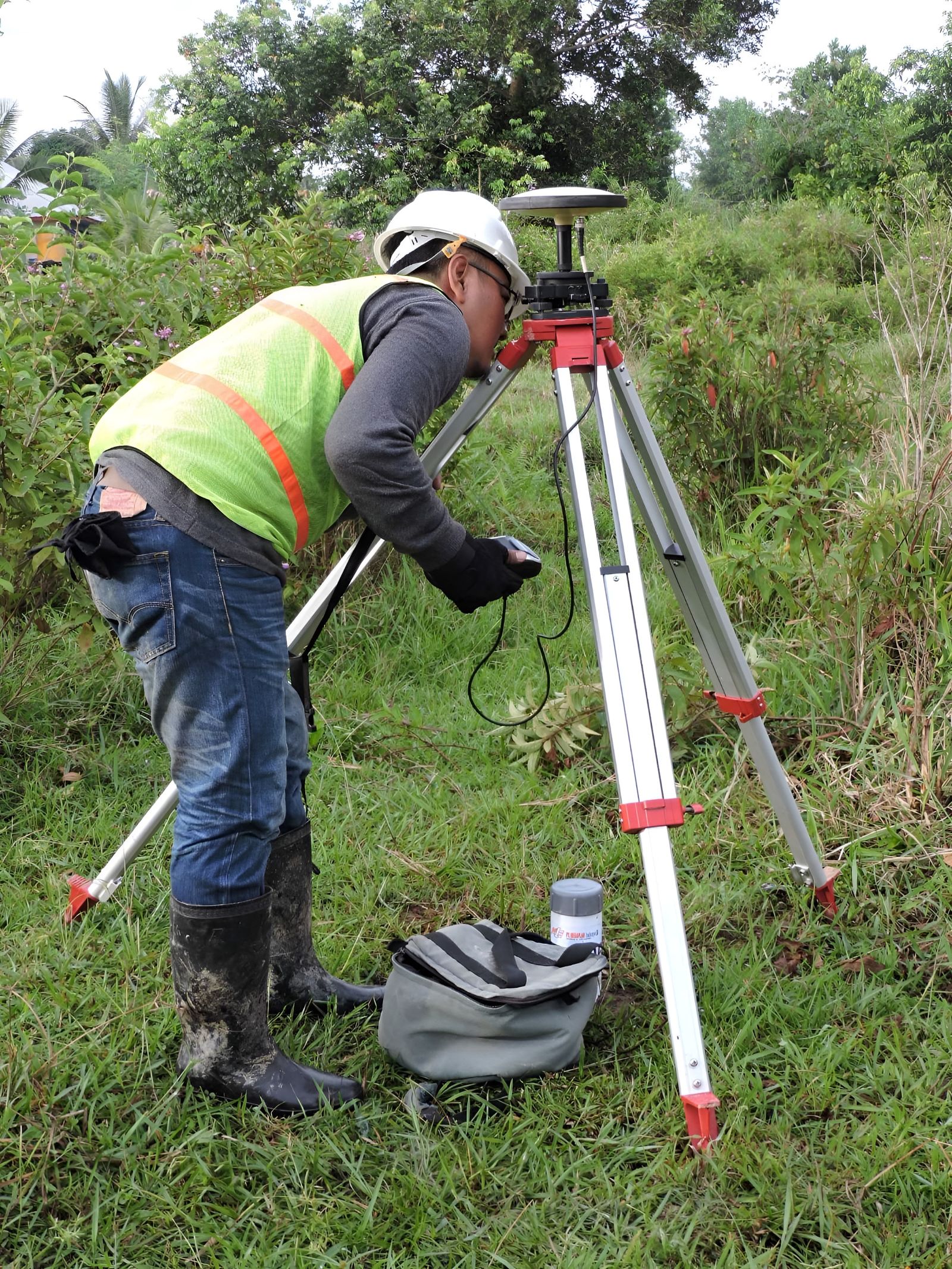 Ilustrasi seorang juru ukur mengoperasikan alat di atas tripod di tengah lahan berumput hijau.