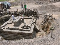 Sisa struktur batu Situs Liyangan yang tersingkap di lahan berkerikil dengan pekerja terlihat di kejauhan.