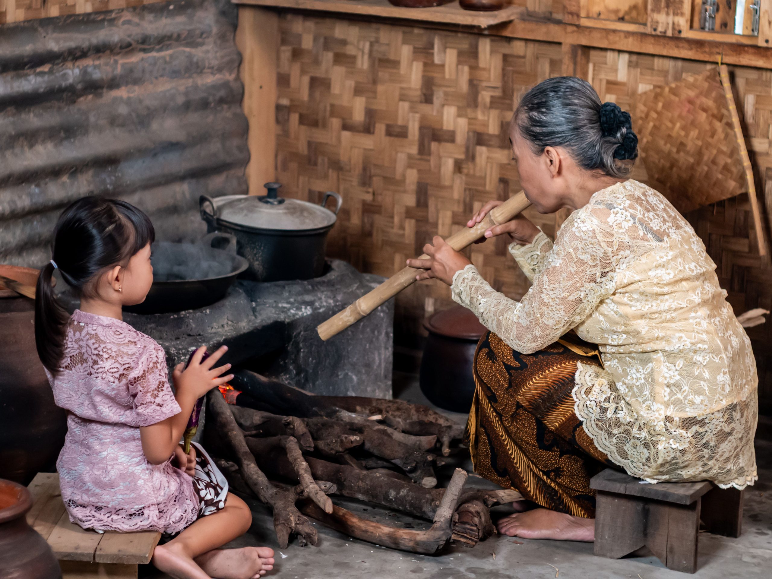 Ilustrasi seorang nenek meniup bara tungku kayu ditemani seorang anak perempuan.