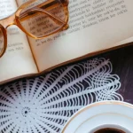 Ilustrasi buku terbuka dengan kacamata baca dan secangkir kopi di atas meja