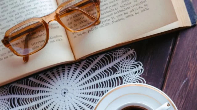 Ilustrasi buku terbuka dengan kacamata baca dan secangkir kopi di atas meja