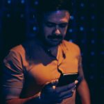Ilustrasi seorang lelaki berkaus jingga menatap layar telepon pintar di ruangan bercahaya kebiruan yang temaram.