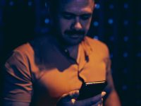 Ilustrasi seorang lelaki berkaus jingga menatap layar telepon pintar di ruangan bercahaya kebiruan yang temaram.