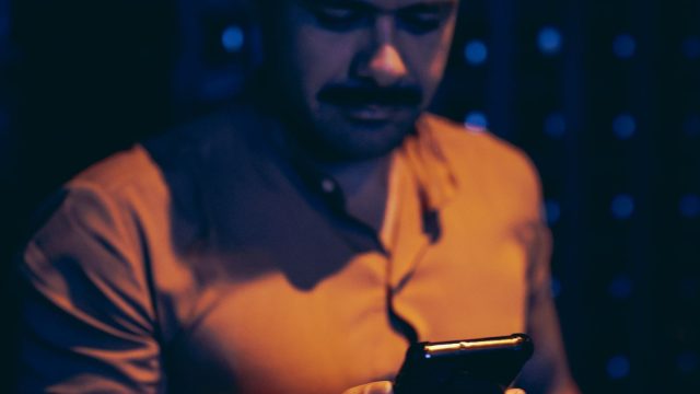 Ilustrasi seorang lelaki berkaus jingga menatap layar telepon pintar di ruangan bercahaya kebiruan yang temaram.