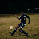 Dua pemain sepak bola berebut bola di lapangan rumput pada malam hari