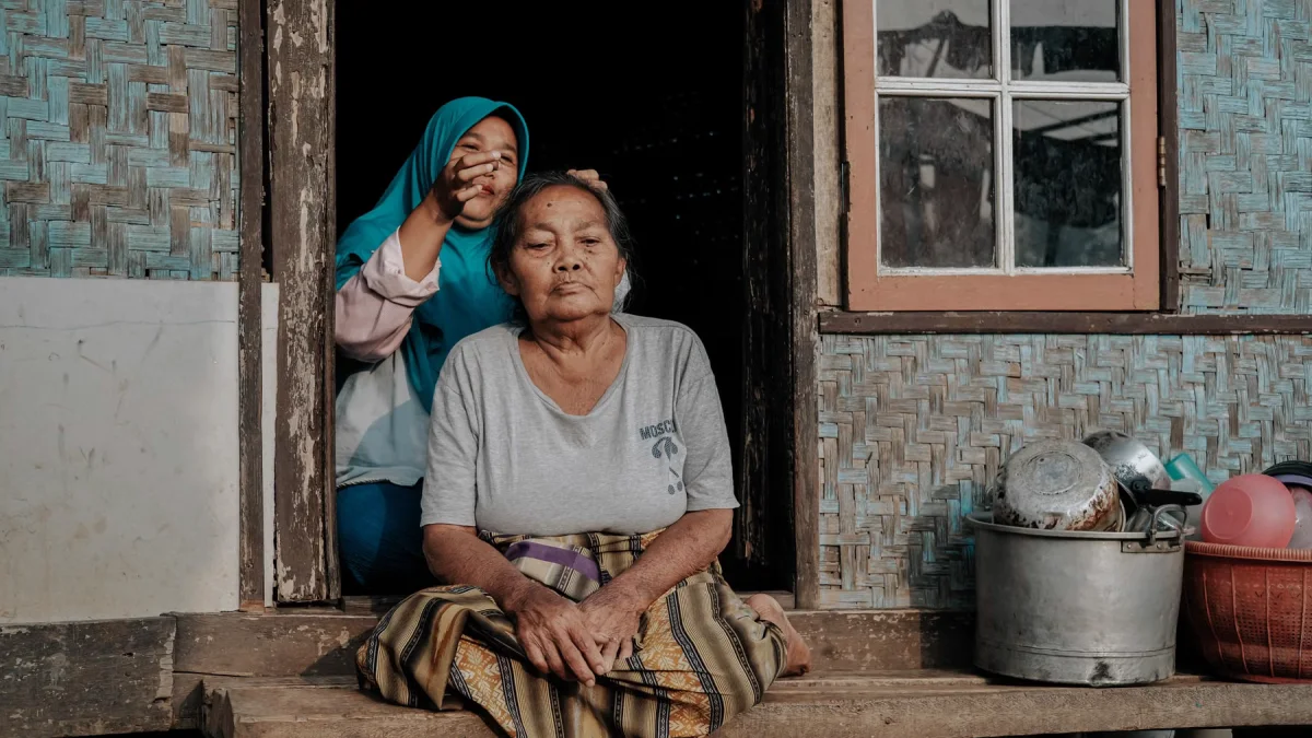Perempuan lanjut usia duduk di teras kayu dengan perempuan muda berkerudung di belakangnya