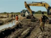Ekskavator kuning dan para pekerja menggali tanah di lahan pedesaan dekat parit berair