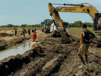Ekskavator kuning dan para pekerja menggali tanah di lahan pedesaan dekat parit berair